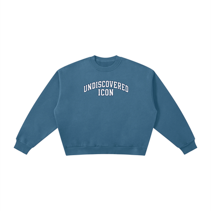 A dusty blue cropped crewneck sweatshirt with 'UNDISCOVERED ICON' printed in white arched block letters across the chest, displayed flat against a solid white background.
