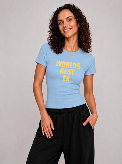 A woman wearing a light blue crewneck t-shirt with yellow text that reads 'WORLDS BEST EX' and black sweatpants, standing against a neutral gray studio background.